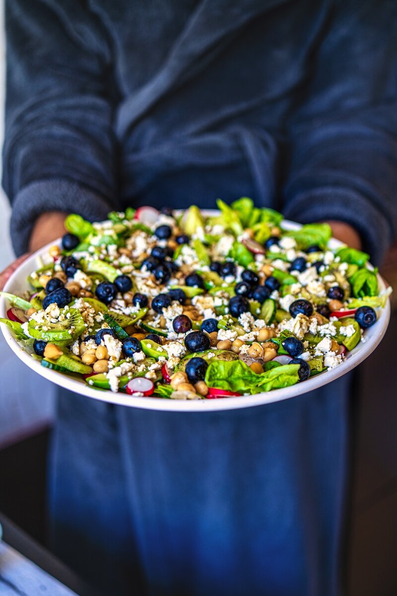 Person holding a large platter of kiwi chickpea feta salad made with avocado, blueberries, chickpeas, feta, and fresh greens.