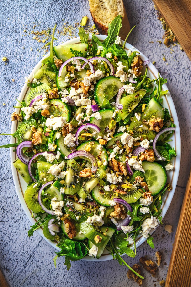 Vibrant green kiwi salad with cucumber slices, arugula, feta, and walnuts on a white plate.