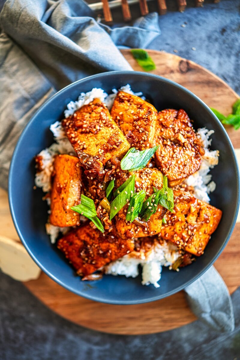 Top view Korean braised tofu rice bowls, hand holding chopsticks, sesame scallion garnish, easy spicy vegan tofu recipe for meal prep