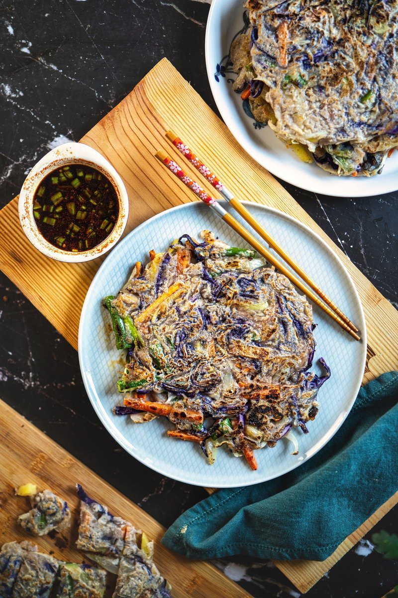 A plate of Korean vegetable pancakes served with dipping sauce and chopsticks on a wooden board.