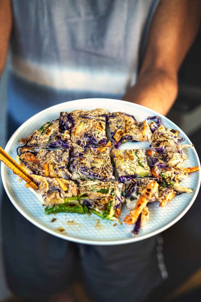 A person holding a plate of Korean vegetable pancakes, featuring colorful vegetables like carrots and scallions.