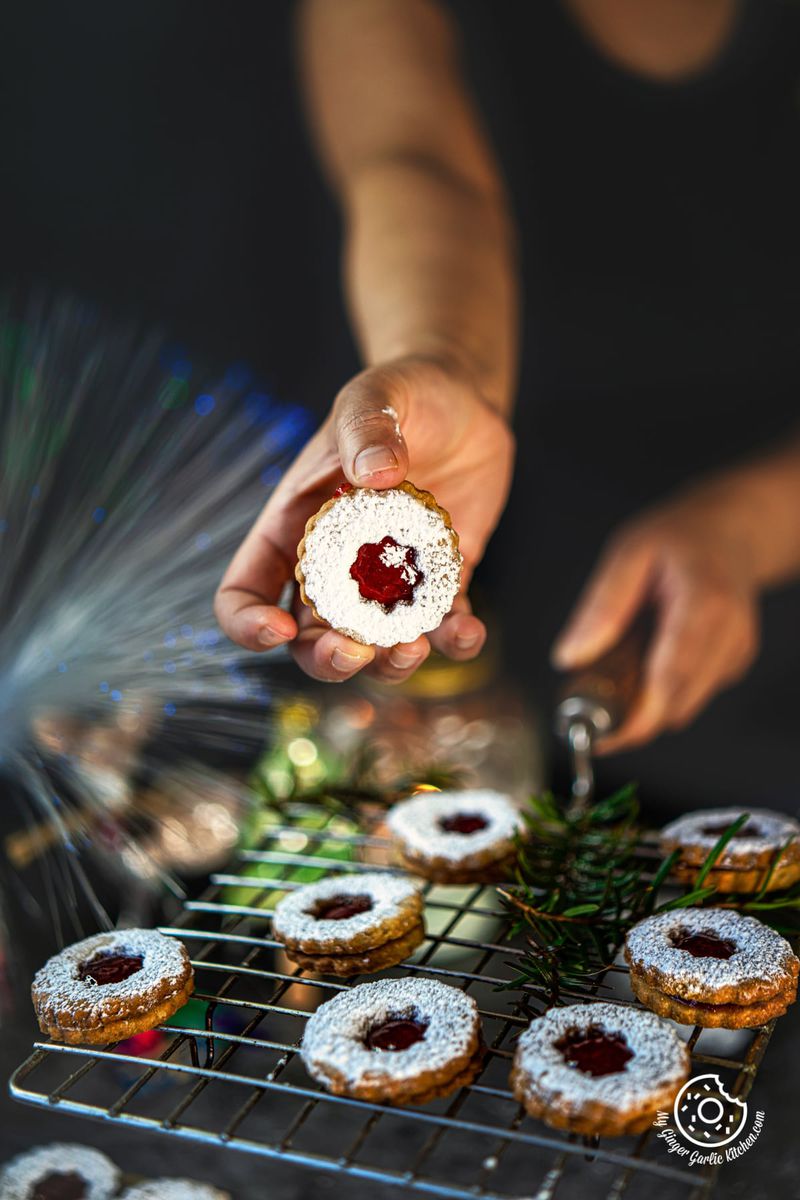 a hand holding 1 linzer cookie with seven linzer cookies on a cooling wrack