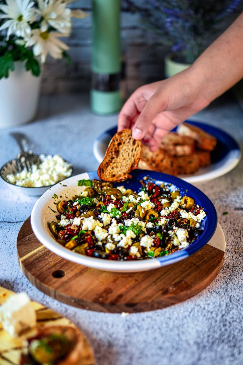 Crispy toasted baguette being dipped into loaded olive oil bread dip with olives, sun-dried tomatoes, feta, and herbs