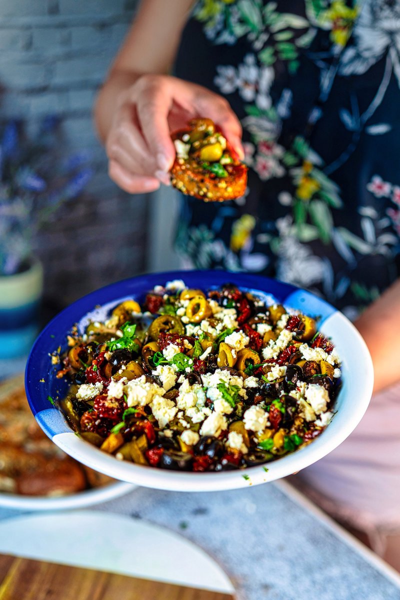 Serving loaded olive oil bread dip with a crispy baguette slice topped with olives, sun-dried tomatoes, feta, and herbs