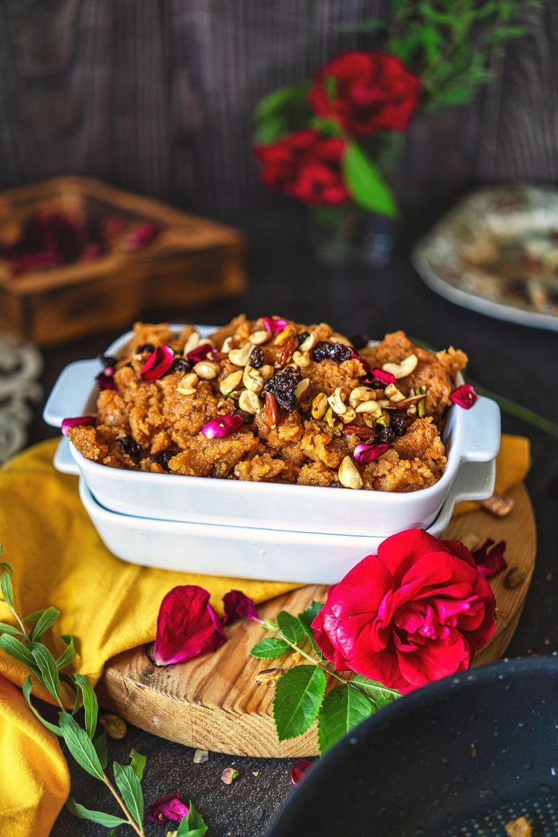 Makhandi halwa served in a white dish, garnished with nuts and rose petals, surrounded by red roses and a yellow fabric for a vibrant contrast.