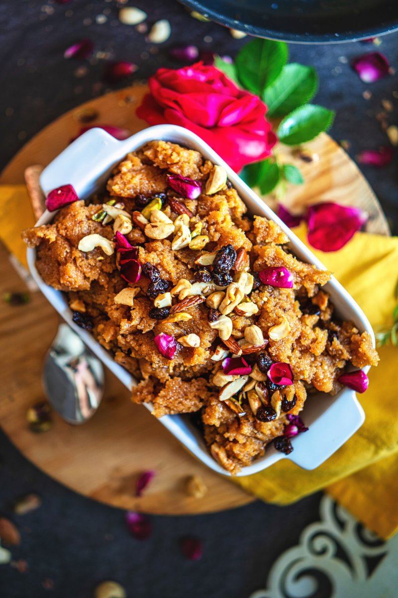 Top view of makhandi halwa in a white dish, topped with assorted nuts and rose petals, set against a dark background with decorative flowers.