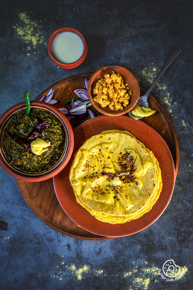 overhead shot of a stack of makki ki roti topped with butter on a brown plate with a bowl of sarson ka saad saag, a bowl of powdered jaggery, a glass of buttermilk and sliced onions on the side