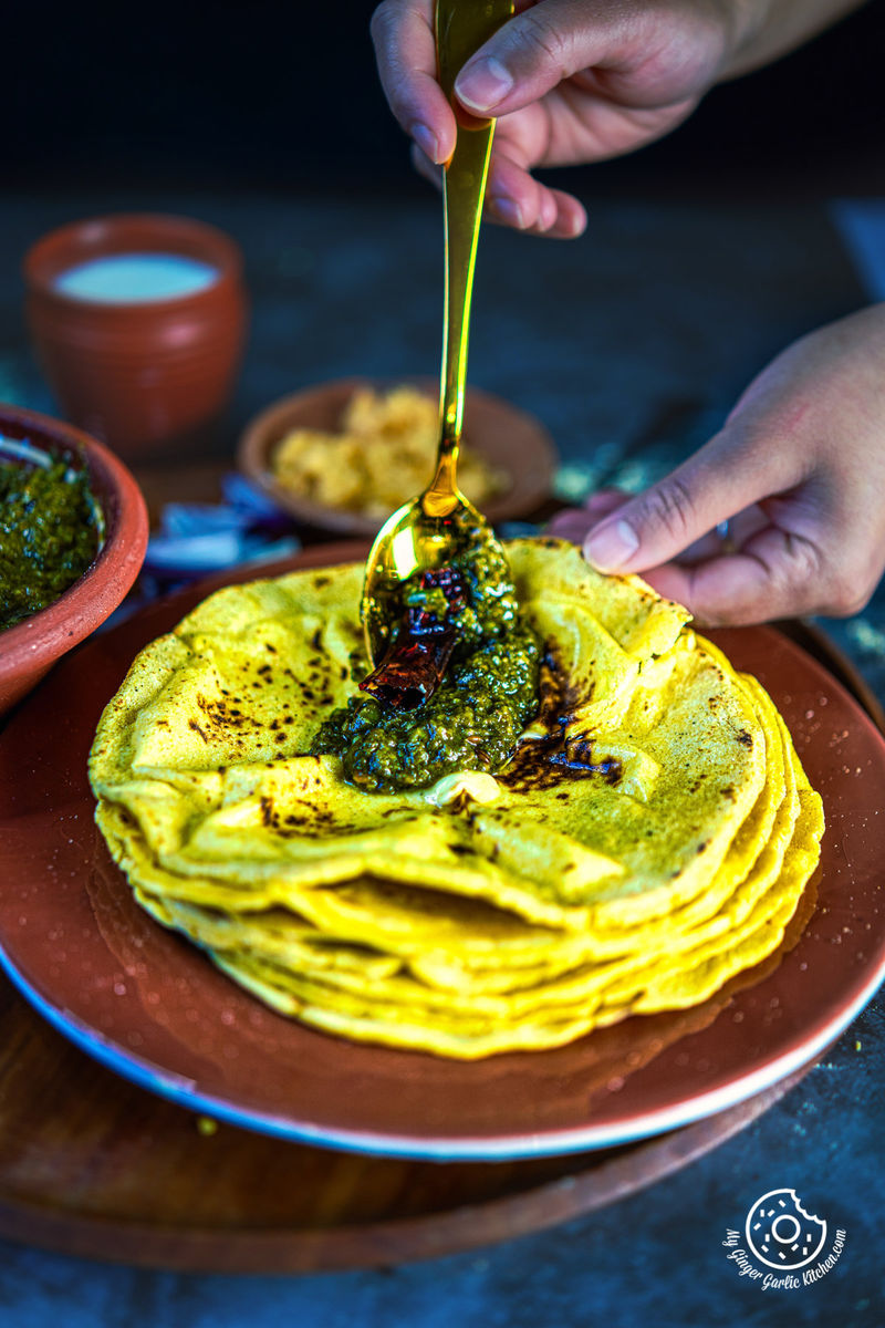 a hand holding a golden spoon with saag over a stack of makki ki roti in brown plate and other hand holding a makki ki roti