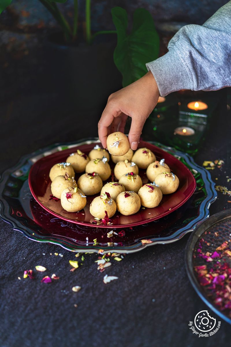 a hand holding malai ladoo over a plate filled with malai laddo