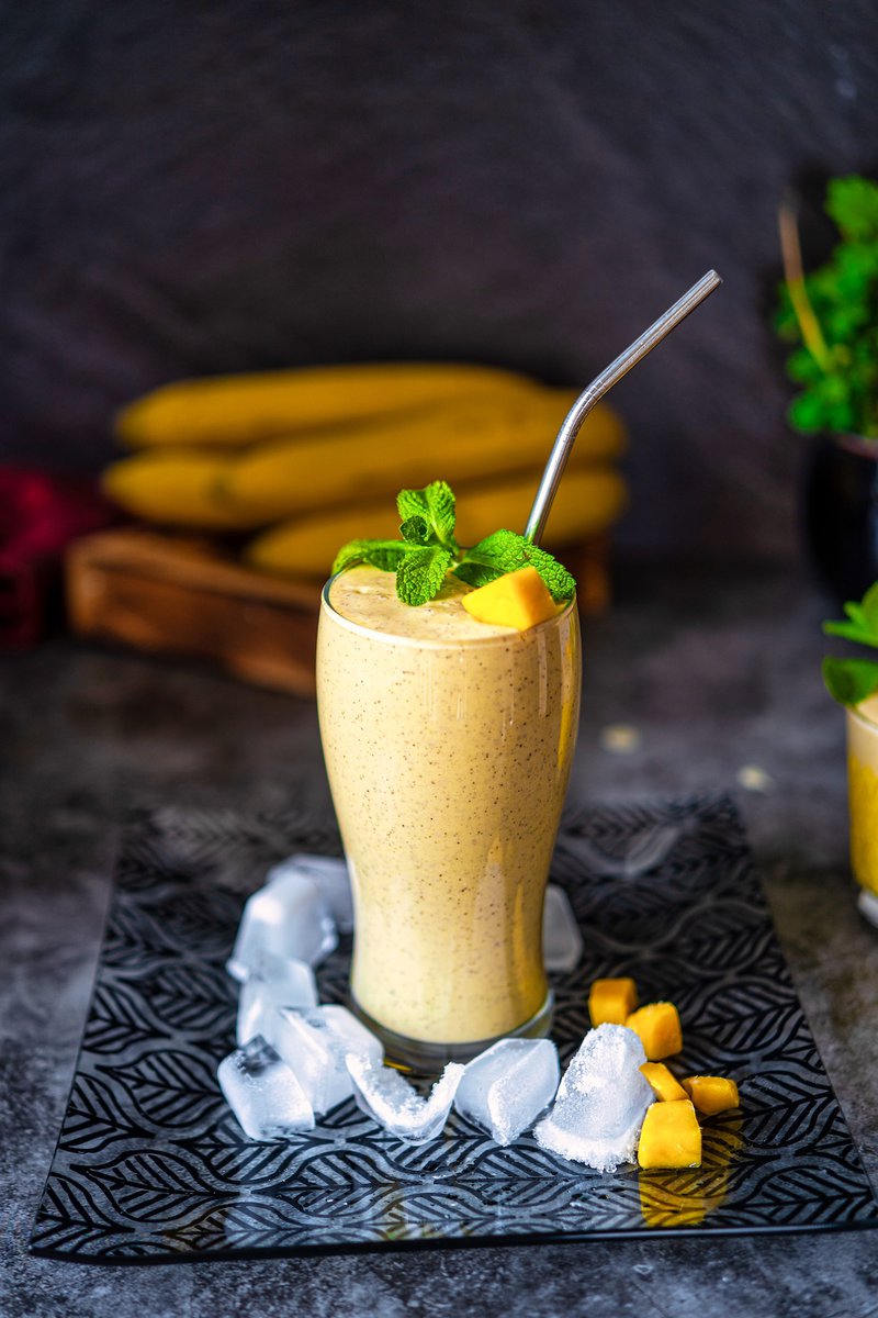 Mango banana smoothie in a tall glass with ice cubes, garnished with mango chunks and fresh mint, on a patterned black mat.