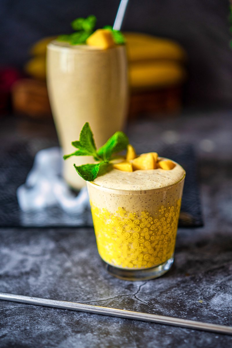 Healthy mango banana smoothie in a decorative yellow glass, garnished with fresh mango and mint on a textured surface.