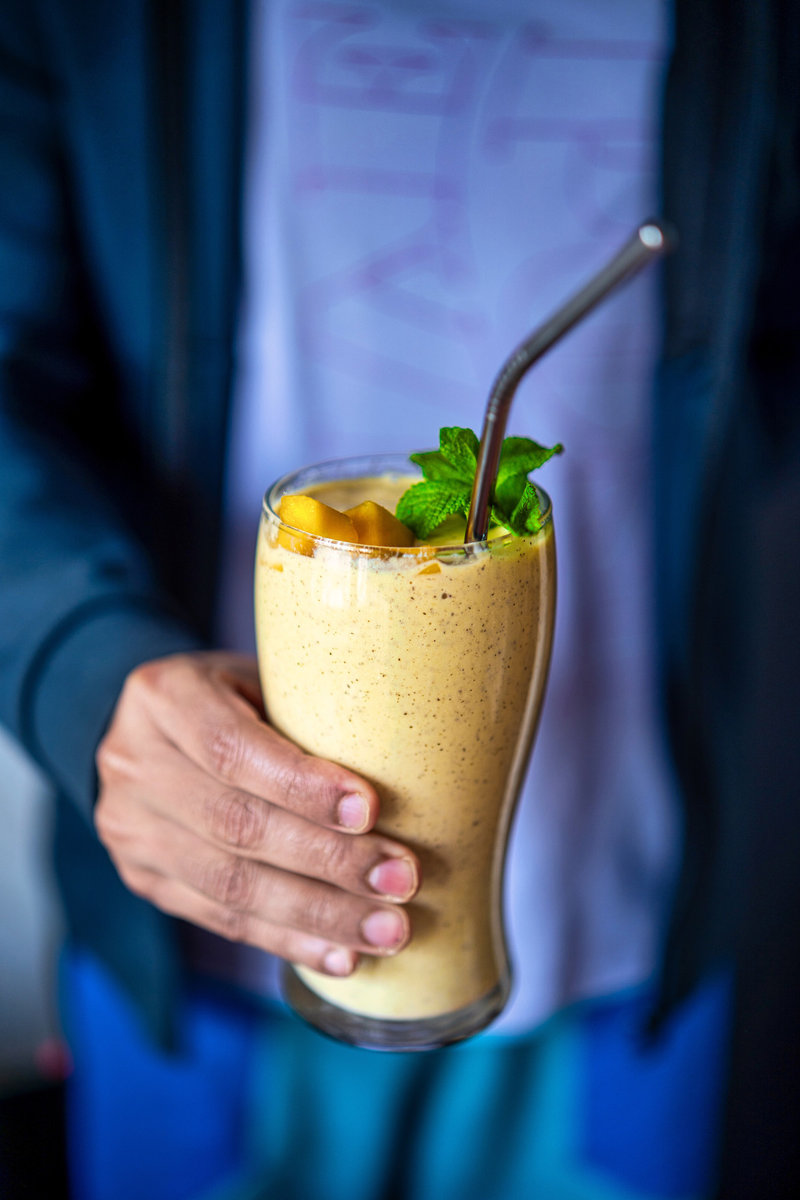 Person holding a mango banana smoothie in a glass, garnished with mango pieces and mint, ready to sip.