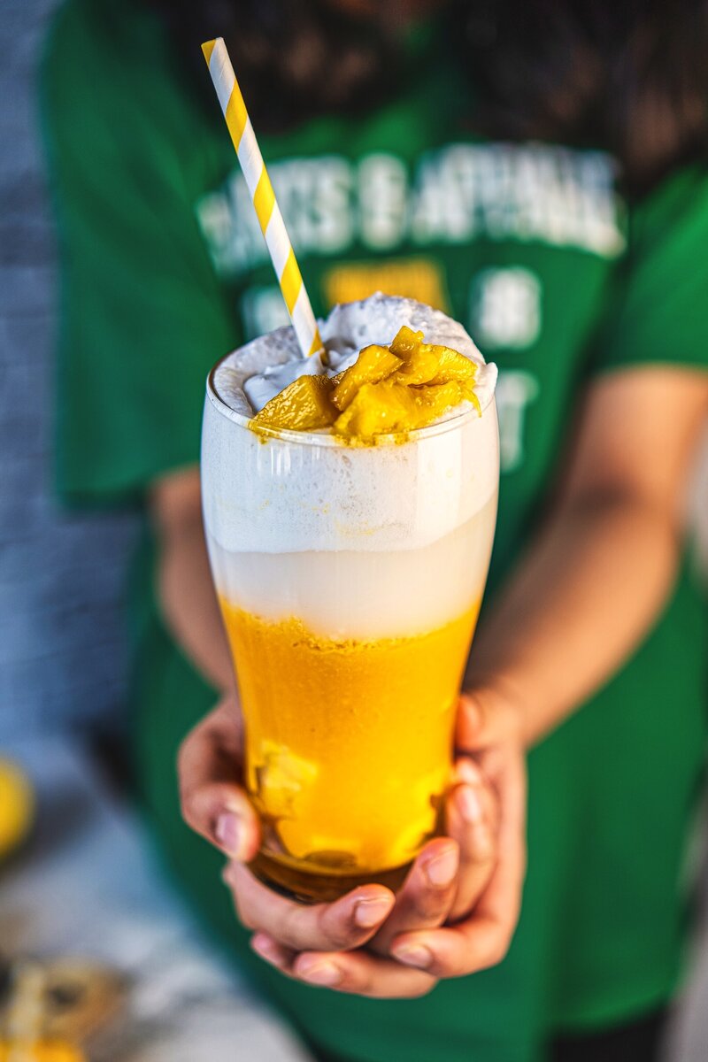 Hand holding mango coconut latte with straw showing creamy layered mango coconut drink, vegan dairy-free beverage
