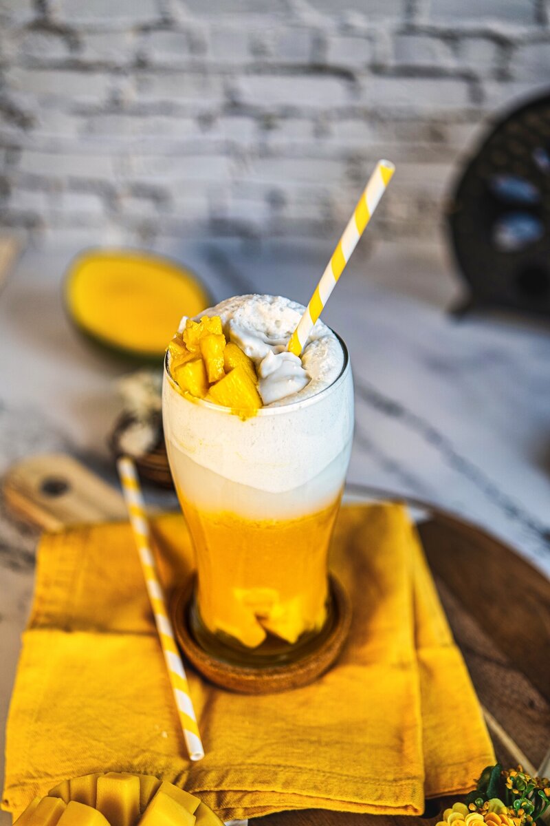 Mango coconut latte styled on yellow cloth with straw and creamy coconut foam topping, tropical mango drink