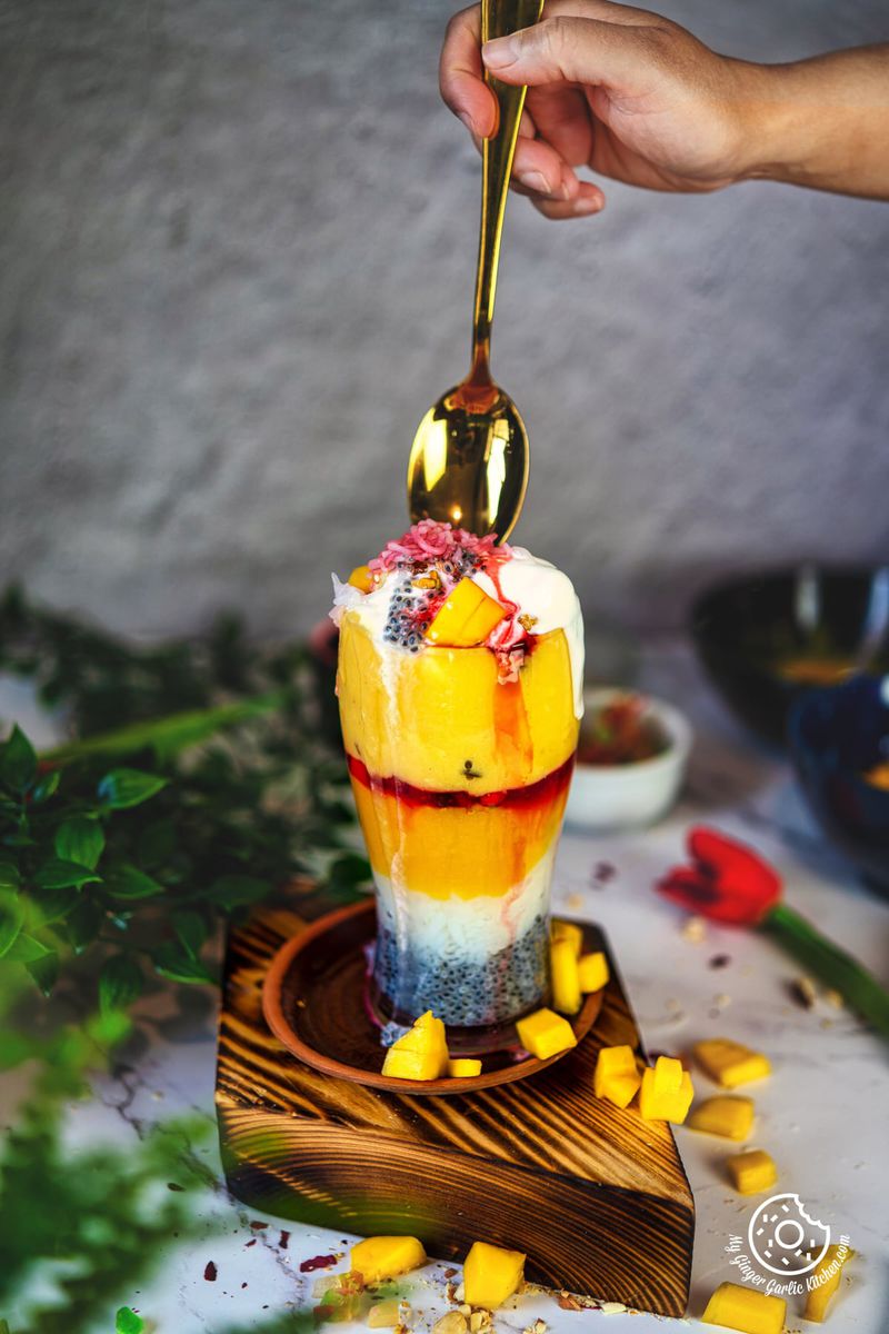 photo of a person is holding a golden spoon over a mango falooda dessert glass