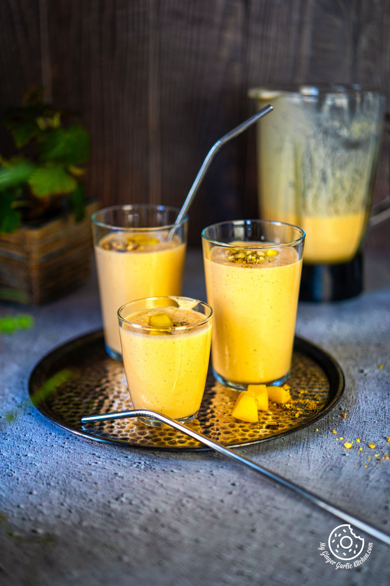 three transparent mango lassi on a metal plate with one steel straw and chopped mangoes