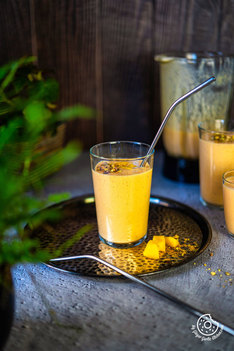 one transparent mango lassi with a steel straw kept on a metal plate with one steel straw and chopped mangoes