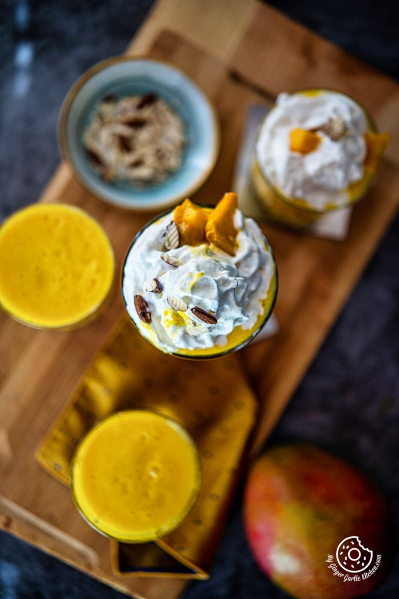 overhead shot of mango milk shake topped with whipped cream and chopped mangoes with more milkshake smoothie glasses on the side