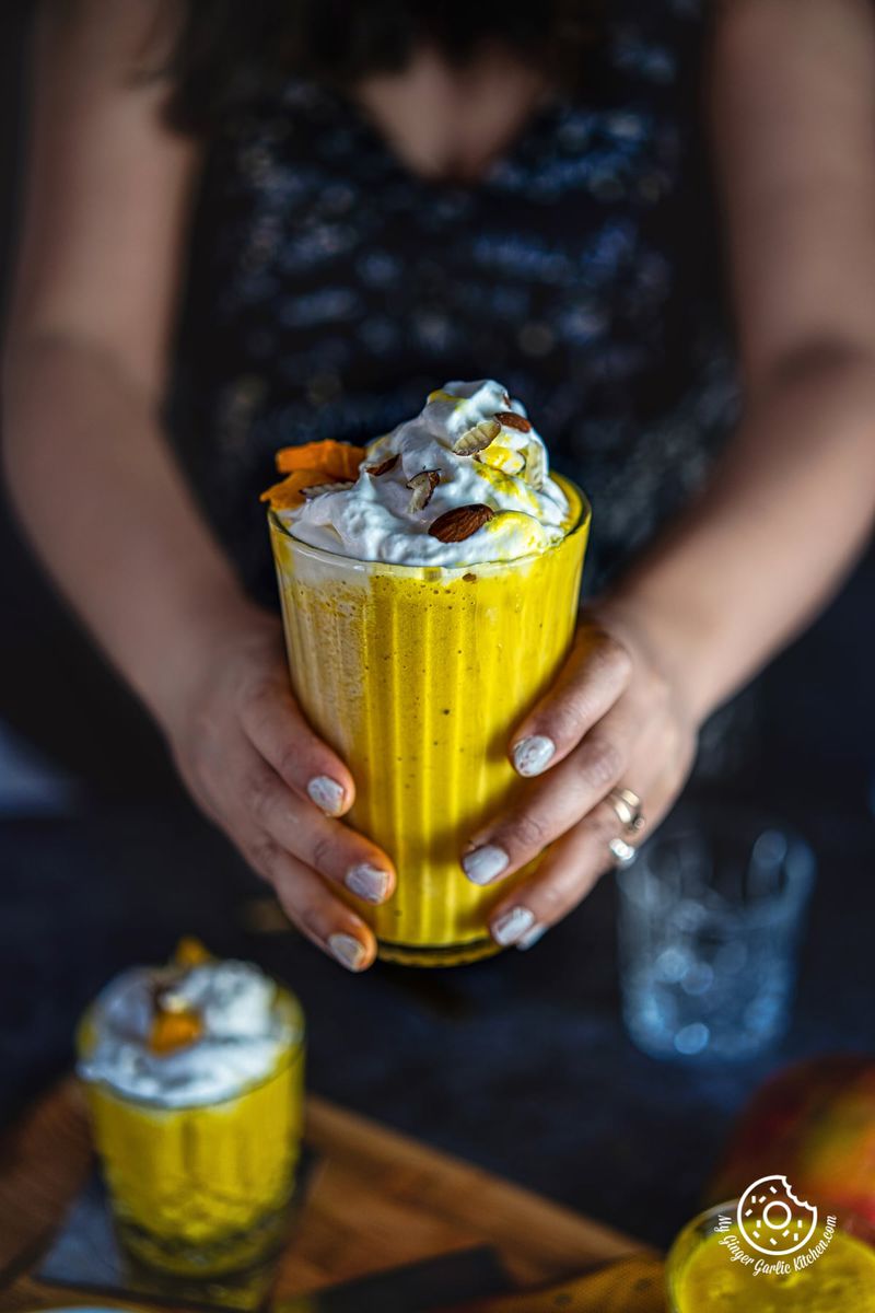 a female holding a tall glass of mango smoothie milkshake topped with whipped cream and nuts