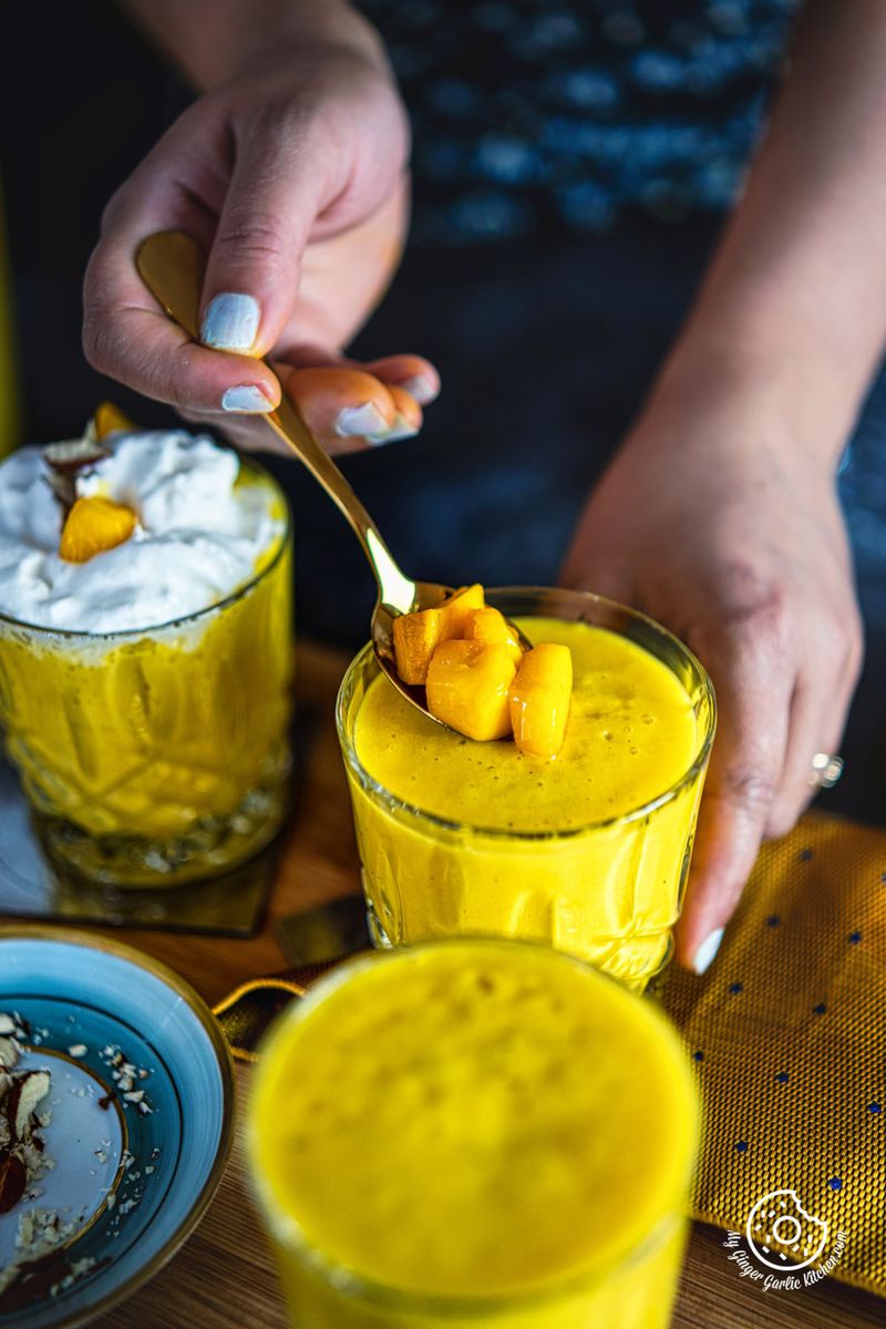 a female hand holding a spoon with chopped mangoes on a mango milkshake glass