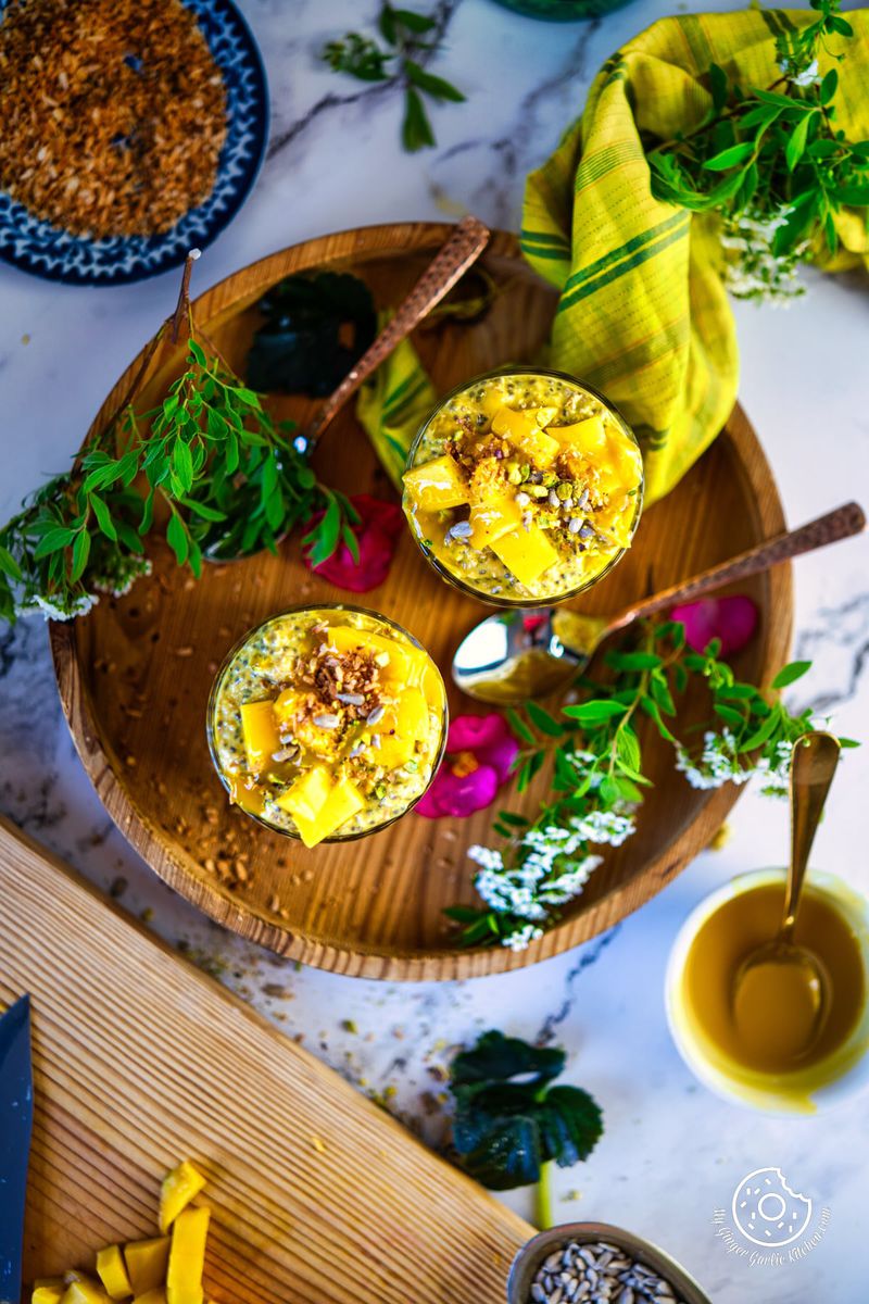 overhead shot of two mango overnight oats glasses on a wooden plate with some fresh flowers