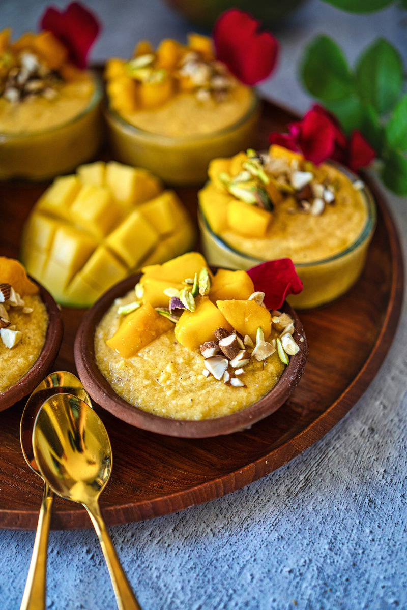 Close-up of mango phirni served in small clay bowls, garnished with diced mangoes, chopped nuts, and rose petals, with golden spoons beside them.