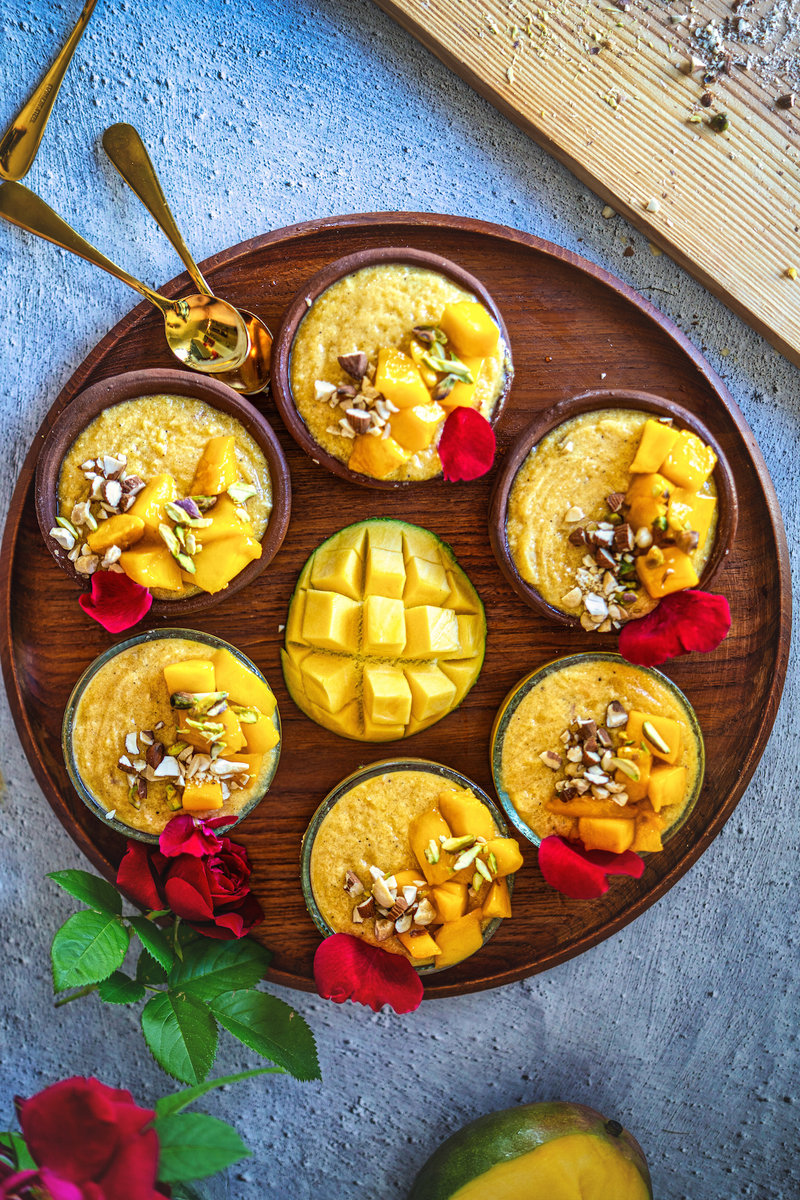 A wooden tray displaying multiple bowls of mango phirni, each topped with fresh mango pieces, nuts, and rose petals, with a beautifully cut mango centerpiece.