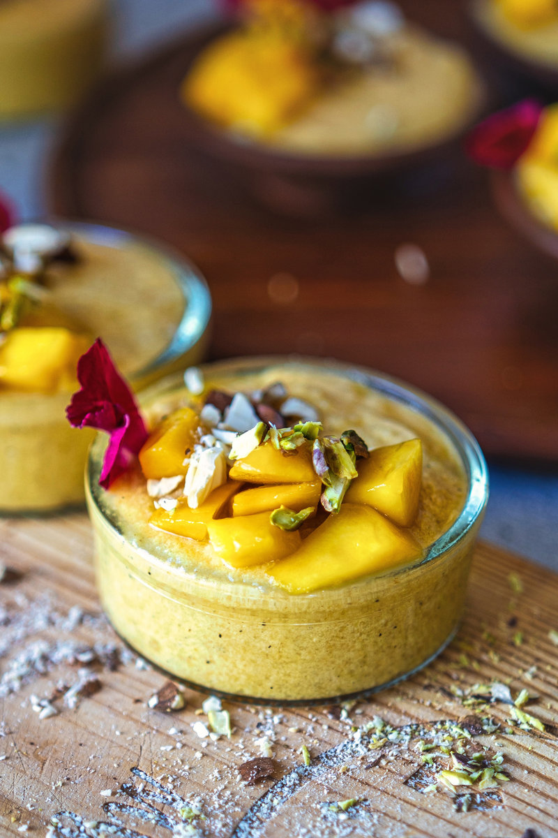 A glass bowl filled with creamy mango phirni, decorated with mango cubes, nuts, and a rose petal, placed on a wooden surface.