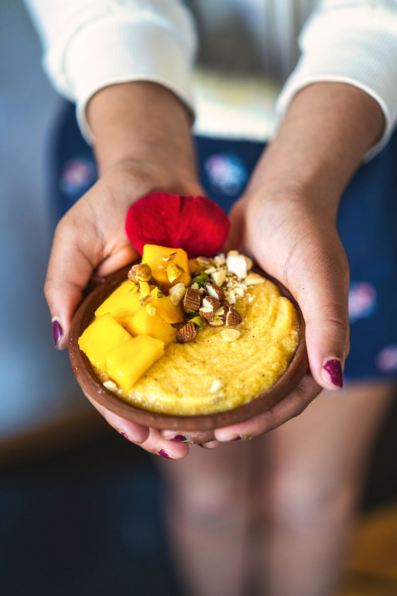 Hands holding a bowl of mango phirni, topped with mango pieces, nuts, and a red rose petal, highlighting the vibrant and delicious dessert.