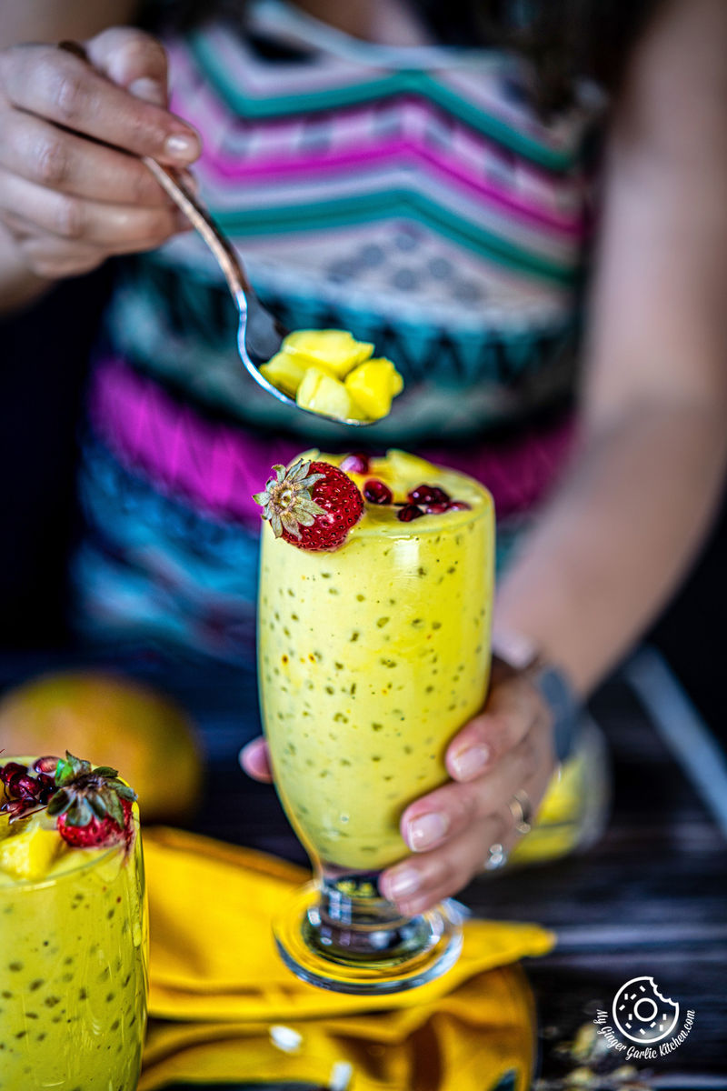 a female holding mango sago glass topped with a fresh mango chunks and strawberry on top and holding a spoon with mango chunks with other hand