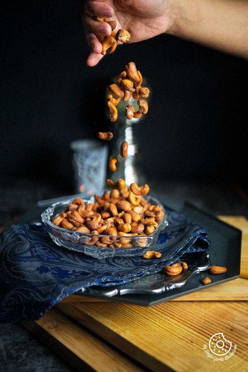 a hand dropping kaju over masala kaju (spicy roasted cashew nuts) bowl