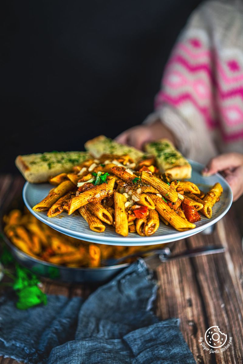 hand holding indian style masala pasta along with garlic bread served in a grey plate