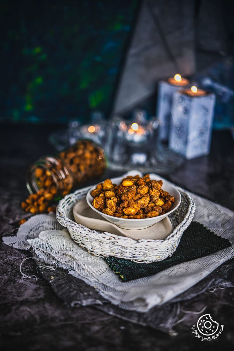 Spiced masala peanuts in a white bowl on a woven mat, with lit candles and a jar of peanuts in the blurred background.