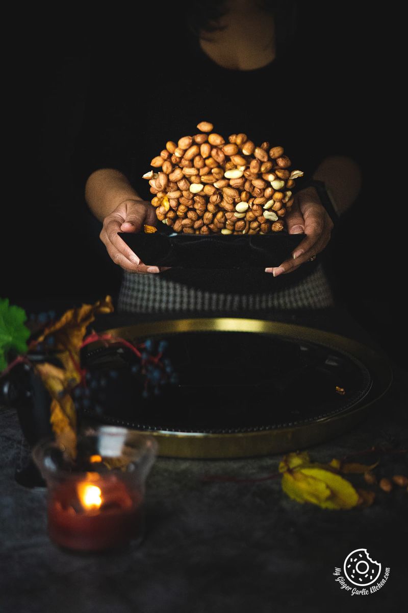 Hands holding a black bowl overflowing with golden masala peanuts, with dark moody lighting and a candle in the background.