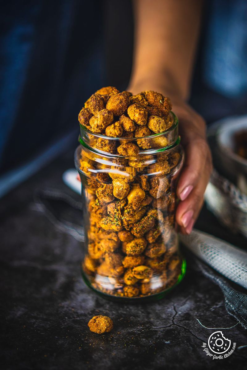 A glass jar full of crunchy masala peanuts held by a person with a dark blue backdrop.