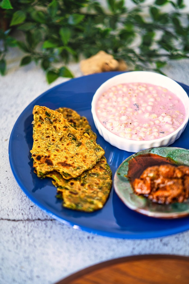 A blue plate with methi parathas, a small bowl of pink yogurt sauce with grains, and a green dish containing a spice blend.