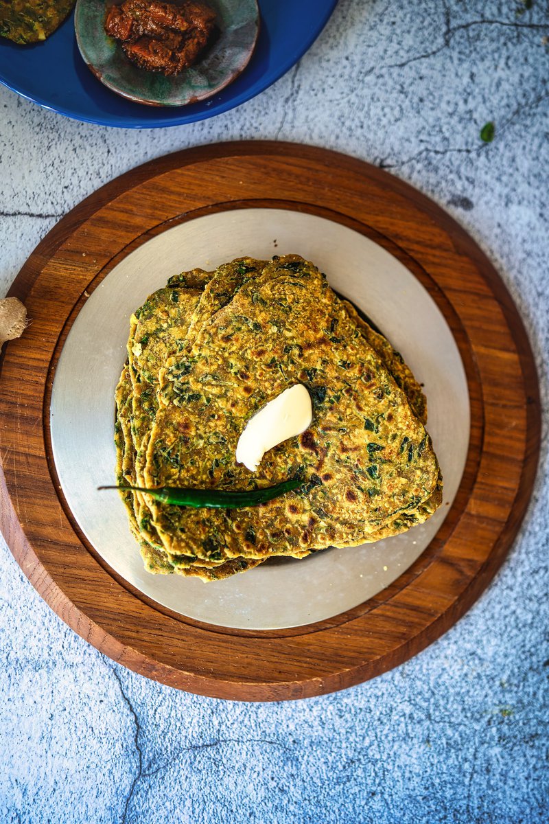 Stack of seasoned fengugreek leaves flatbreads aka methi paratha with herbs, topped with a dollop of butter and a green chili, on a wooden plate.