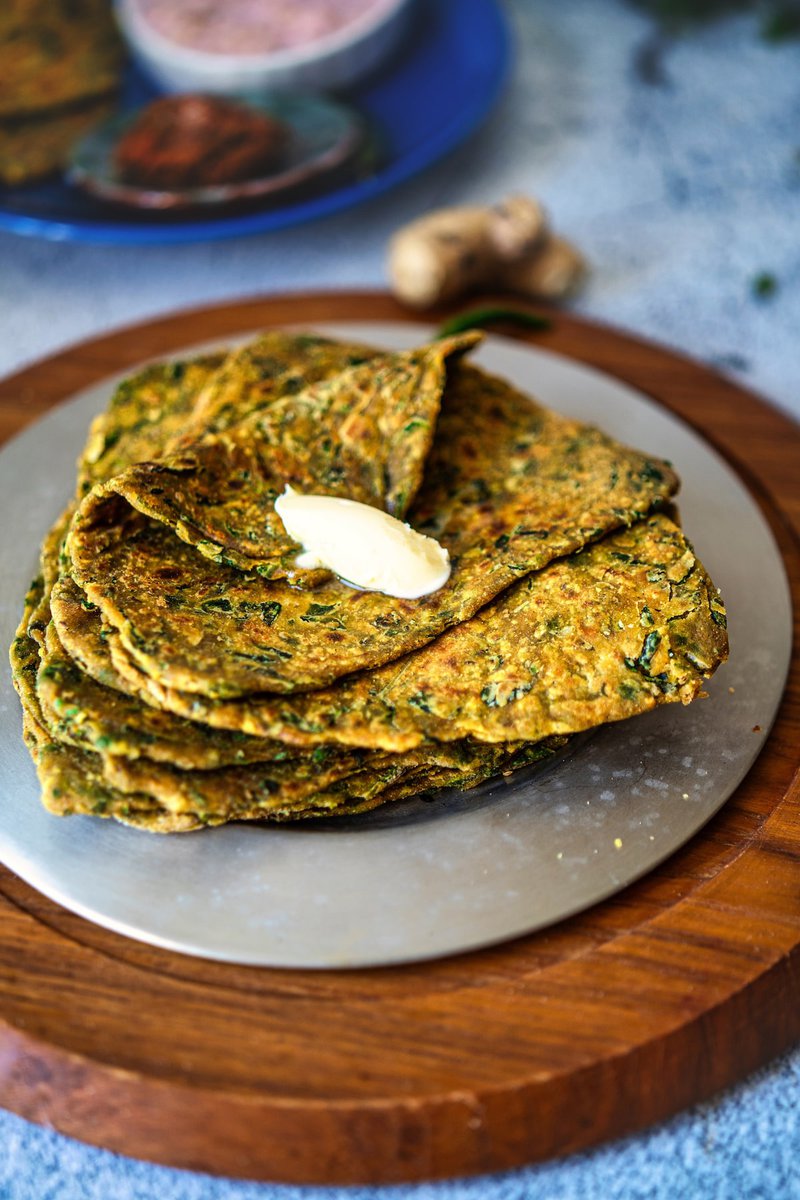 A stack of methi paratha flatbreads served on a round wooden board with a dollop of butter on top.