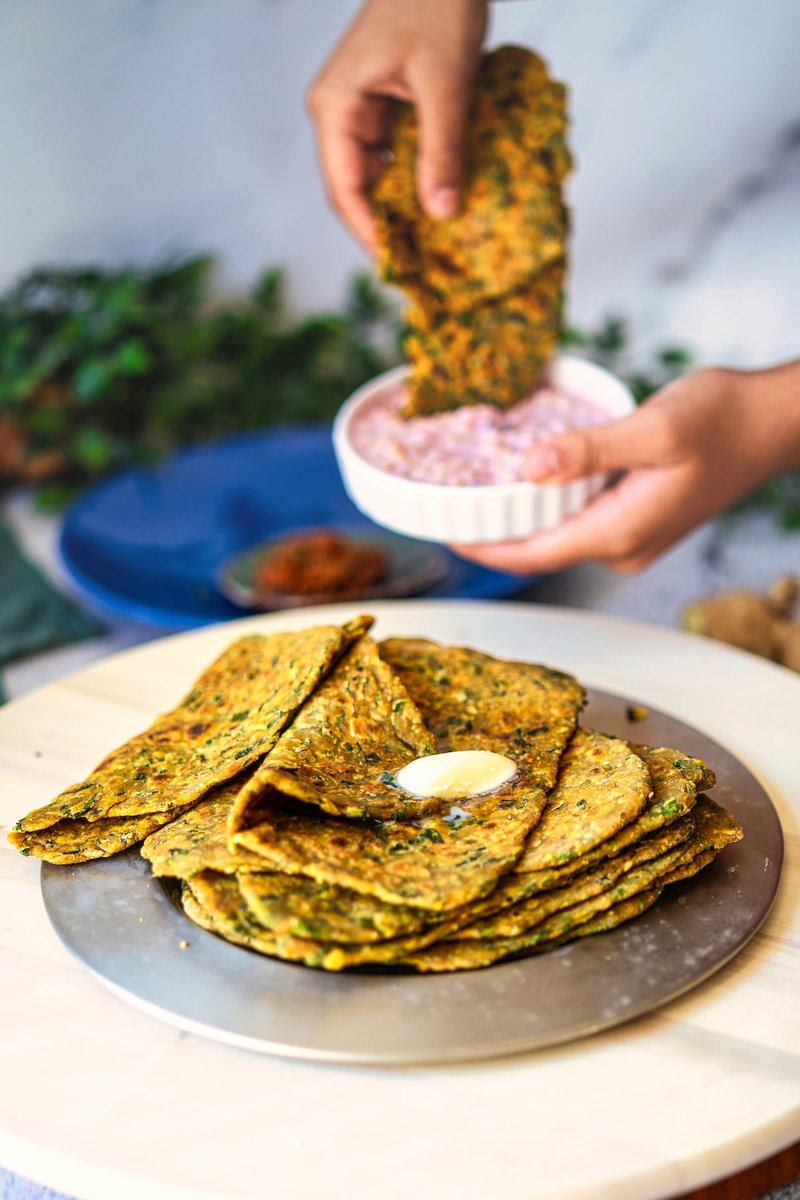 A stack of spiced methi parathas on a plate with a person dipping one into a bowl of chutney.