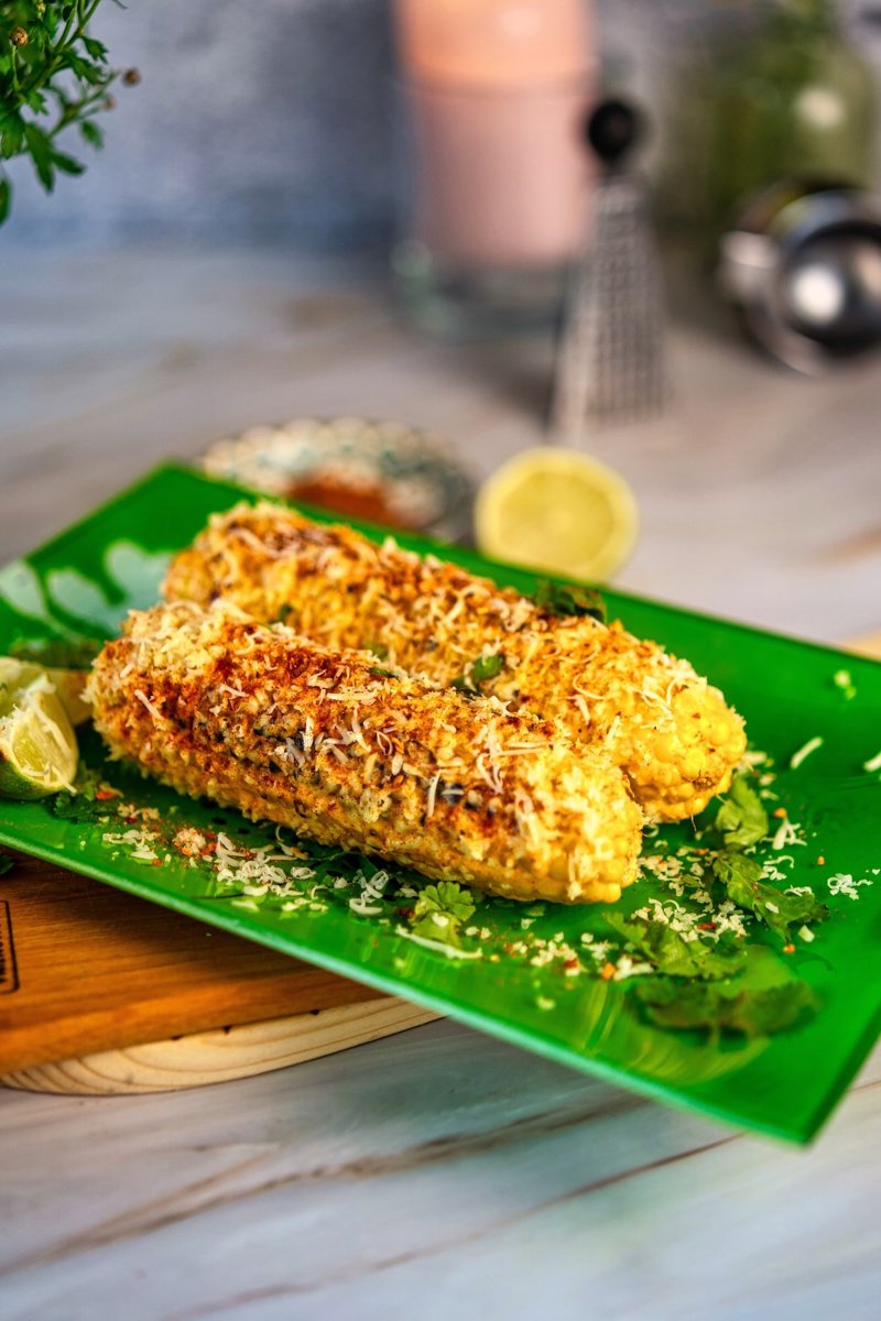 Two grilled Mexican street corn elotes topped with creamy sauce, grated cheese, chili seasoning, cilantro, and lime