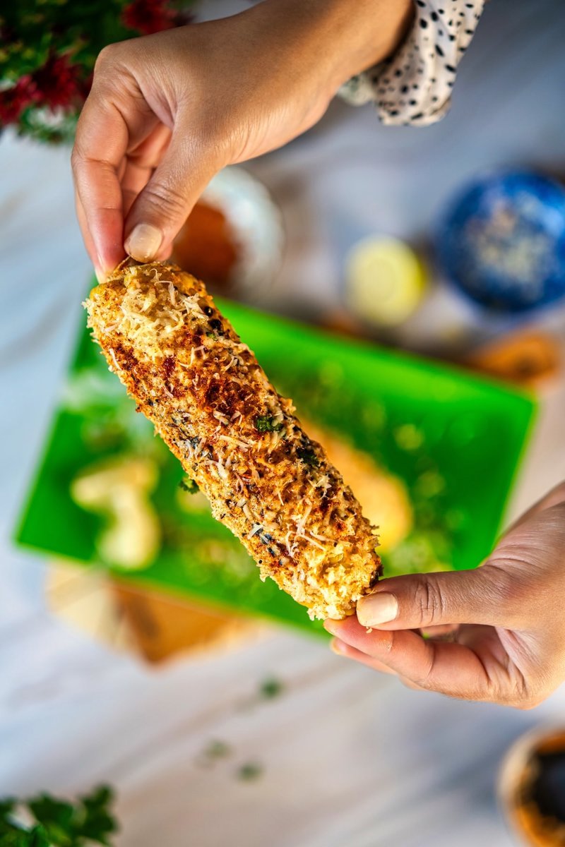 Mexican street corn elote held in hand, topped with creamy garlic-lime sauce, grated cheese, chili, and cilantro