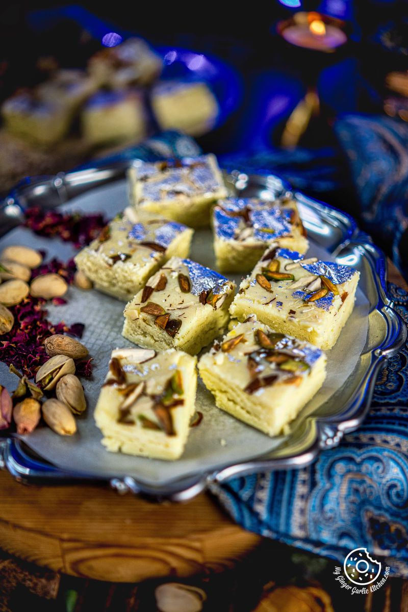 milk barfis in a metal plate with some pistachios and rose petals on the side