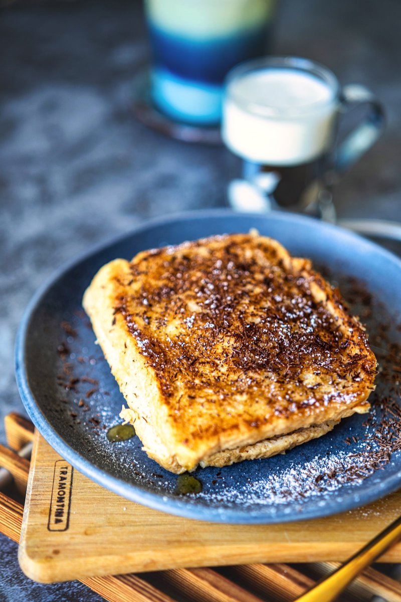 Close-up of a golden-brown milk toast drizzled with honey, sprinkled with dark chocolate shavings and icing sugar, served on a blue plate.