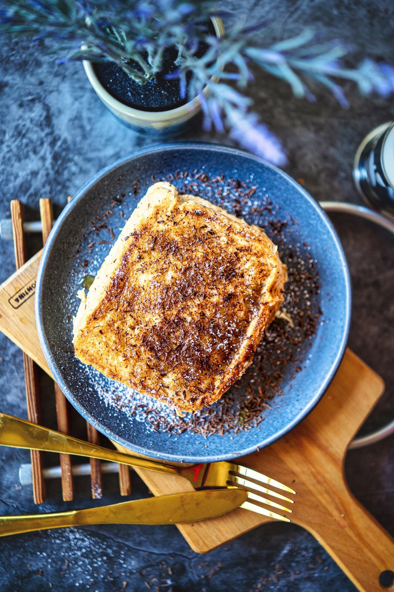 Top view of a decadent milk toast garnished with chocolate shavings and icing sugar, presented on a blue plate with golden cutlery.