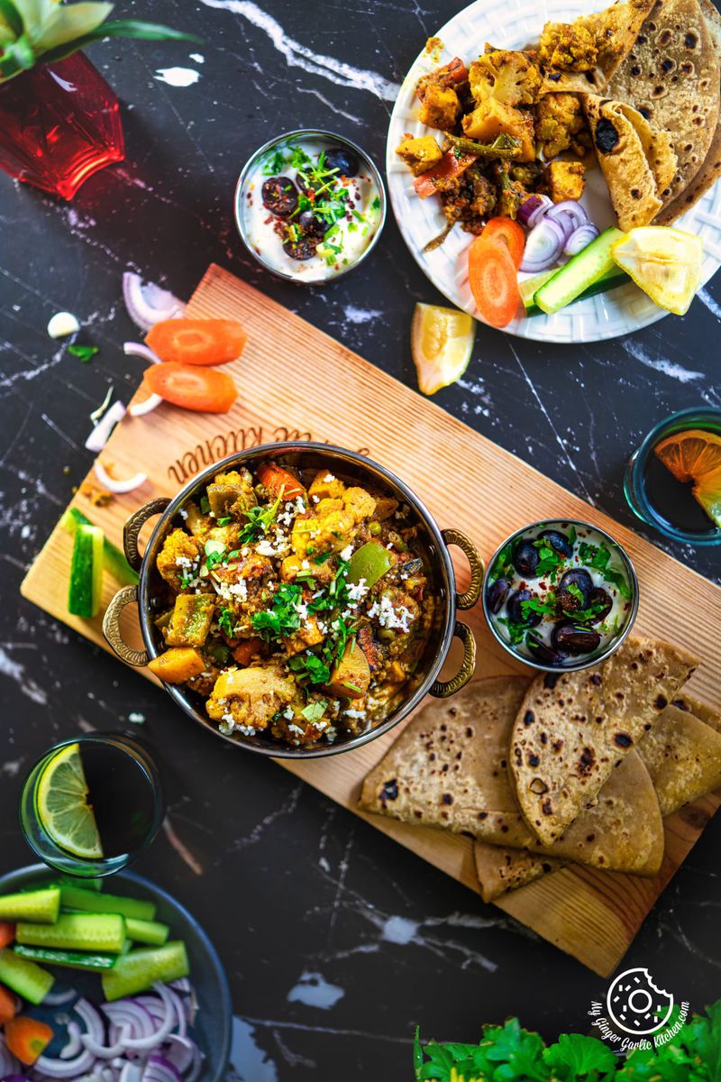 overhead shot of mixed vegetable garnished with coriander on a wooden board with raita and parathas on the side