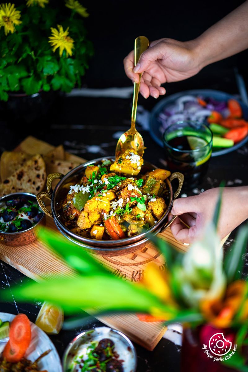 a hand holding a golden spoon over mix veg copper kadai