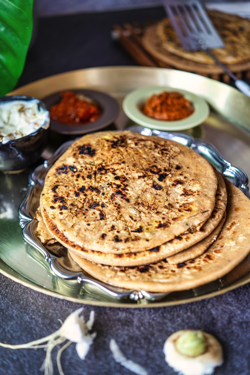 Stack of golden-brown mooli parathas served on a silver platter with bowls of condiments.