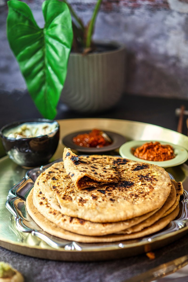 A plate of stacked Mooli Parathas (Radish Parathas) with a piece partially folded, accompanied by bowls of chutneys and yogurt.