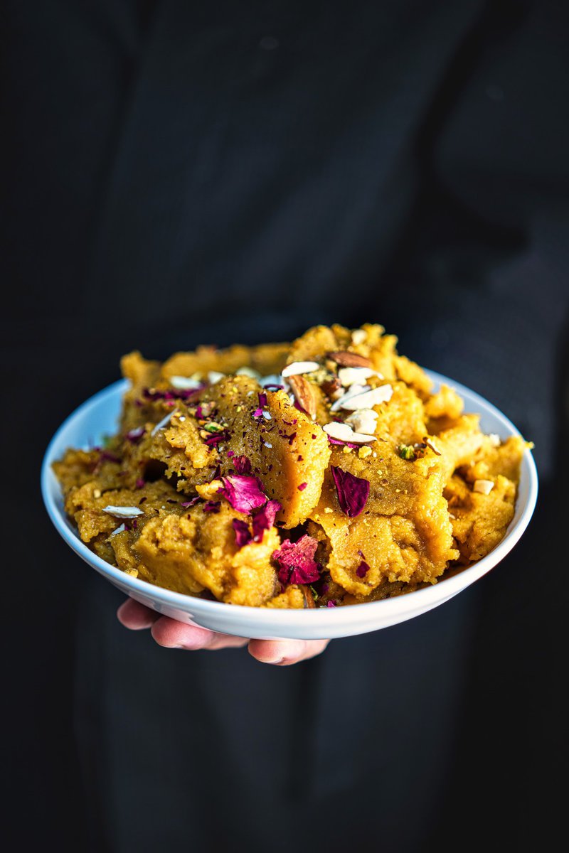 A close-up of a bowl of moong dal halwa, garnished with sliced almonds and dried rose petals, held by a person against a dark background.