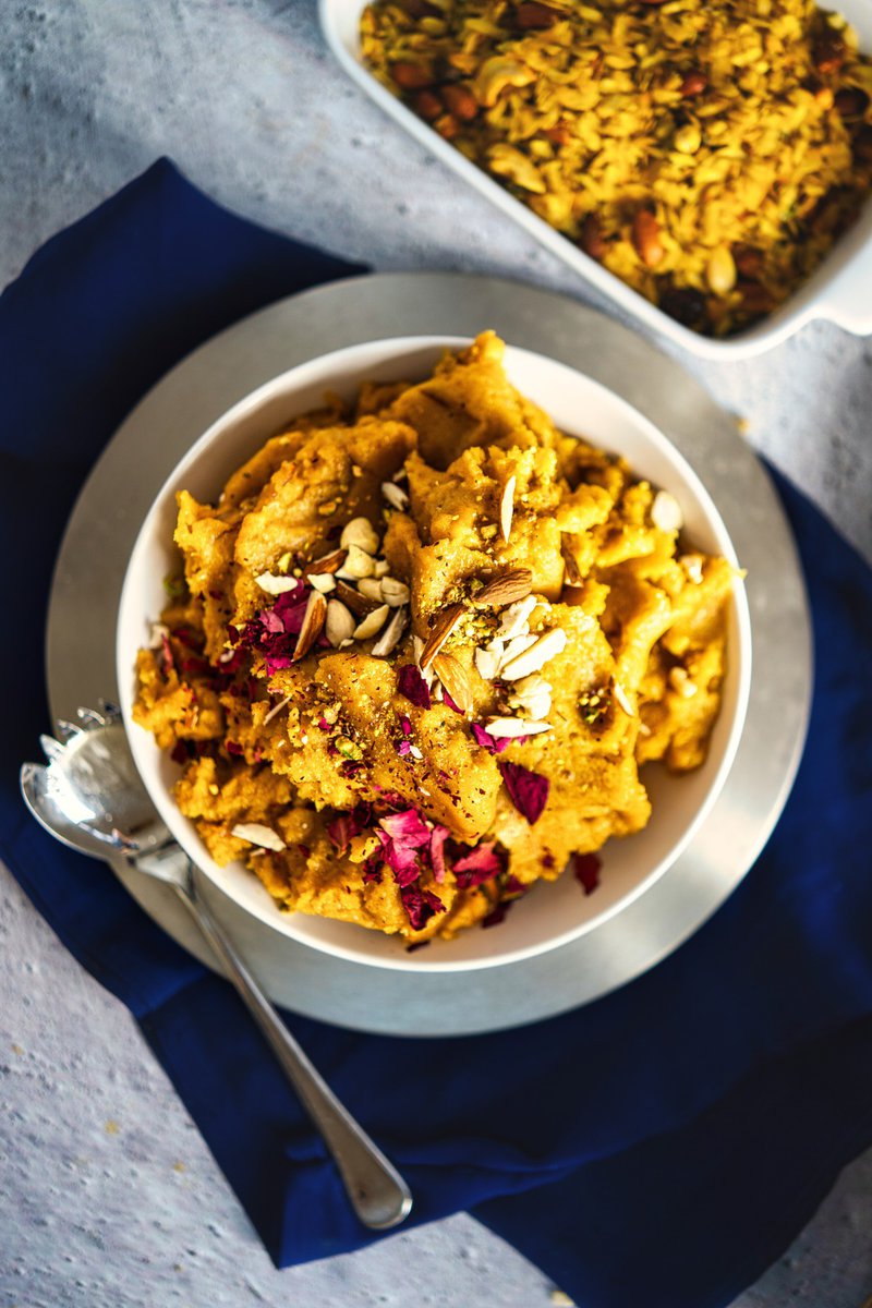 A top view of a bowl of moong dal halwa on a plate with a spoon, garnished with almonds and rose petals, placed on a blue cloth. A serving dish with more halwa is visible in the background.